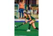 A female field hockey player in a green uniform preparing to hit the ball on a green pitch. A person in an orange vest watches in the background.