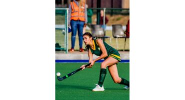 A female field hockey player in a green uniform preparing to hit the ball on a green pitch. A person in an orange vest watches in the background.