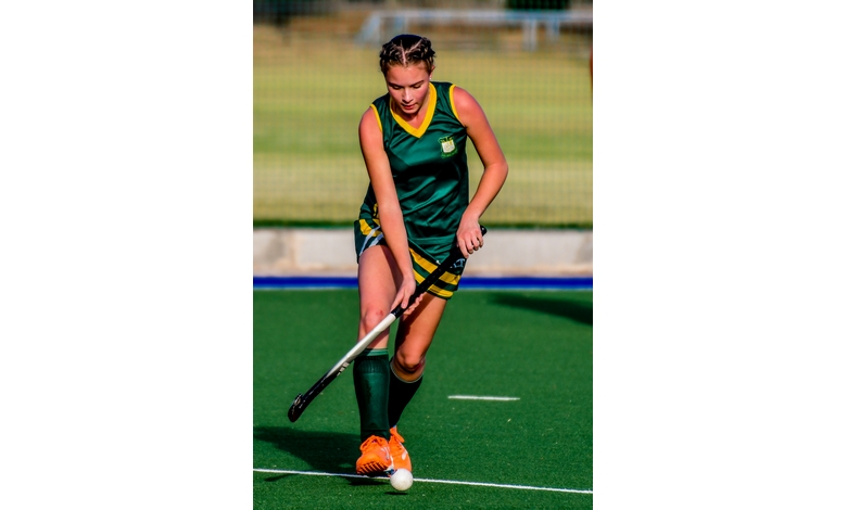 A female field hockey player in green and yellow gear skillfully dribbles a ball on a green turf field, exuding focus and determination.