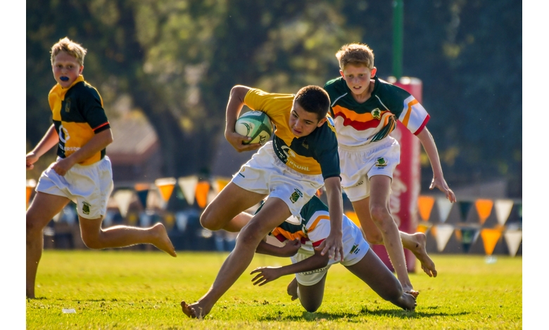 A youth rugby player in a yellow and green jersey evades opponents on a grassy field. Intense action and concentration are captured under bright sunlight.
