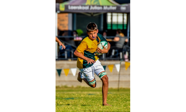 A young boy playing rugby sprints forward determinedly on a grassy field, clutching the ball. Spectators and flag bunting are visible in the background.