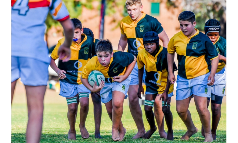 Young rugby players in green and yellow uniforms prepare for a play on a grassy field. A central player holds the ball, focused and ready to advance.