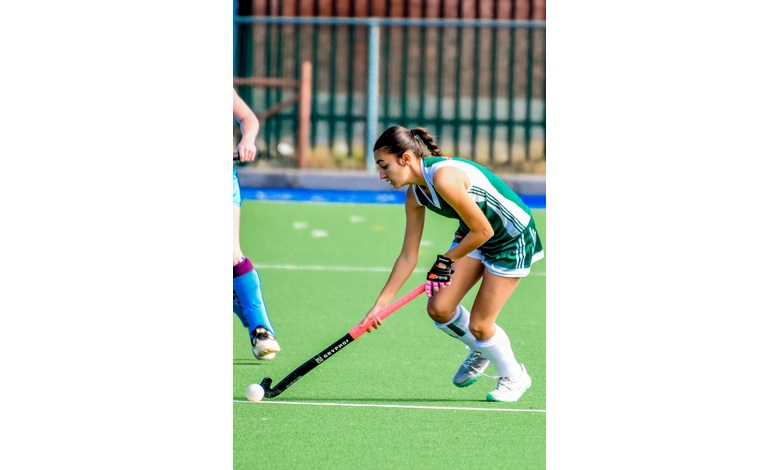 A field hockey player in a green and white uniform is bent forward, skillfully dribbling the ball with a black stick on a green turf. The scene conveys focus and athleticism.
