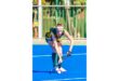 A field hockey player in green and yellow uniform focuses intently, poised to strike a white ball on a bright blue field, conveying determination.