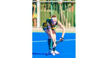 A field hockey player in green and yellow uniform focuses intently, poised to strike a white ball on a bright blue field, conveying determination.
