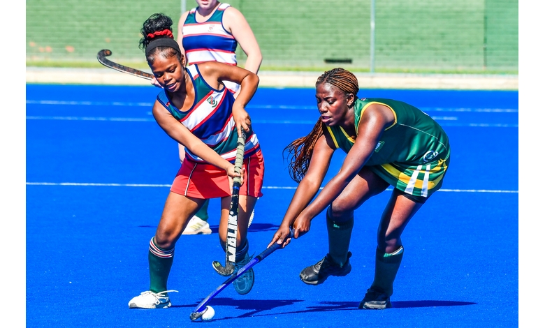 Two female field hockey players in action on a bright blue turf. One player in a red and white striped jersey competes for the ball against another in green and yellow. Intense focus and athleticism are evident.