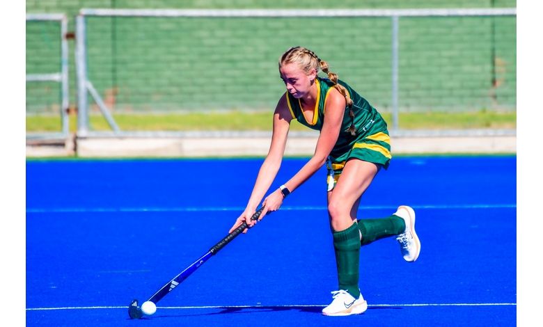 A field hockey player in a green and yellow uniform focuses intently while striking the ball on a bright blue field. Background shows a green wall and fence.