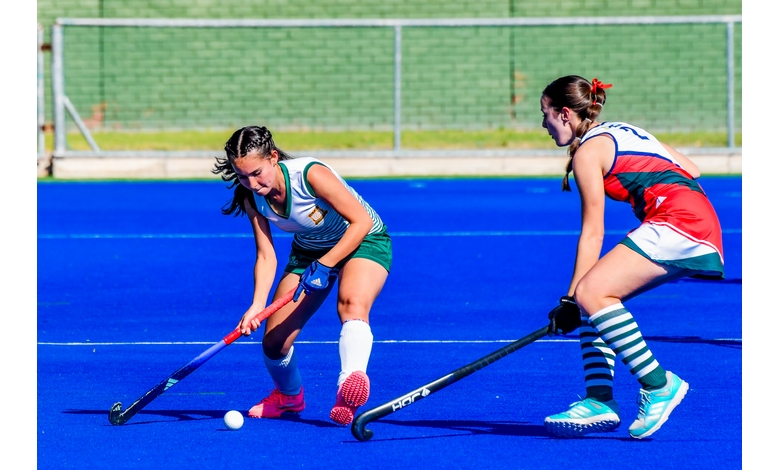 Two female field hockey players compete on a vibrant blue field. One player in green controls the ball, while the other in red defends intensely.