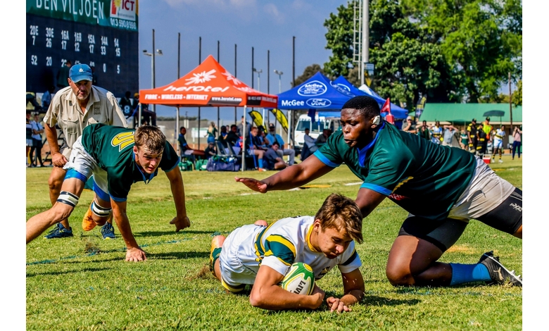 Rugby match action: two players in green jerseys chase a player in a white jersey scoring a try. Onlookers and tents in the vibrant background.