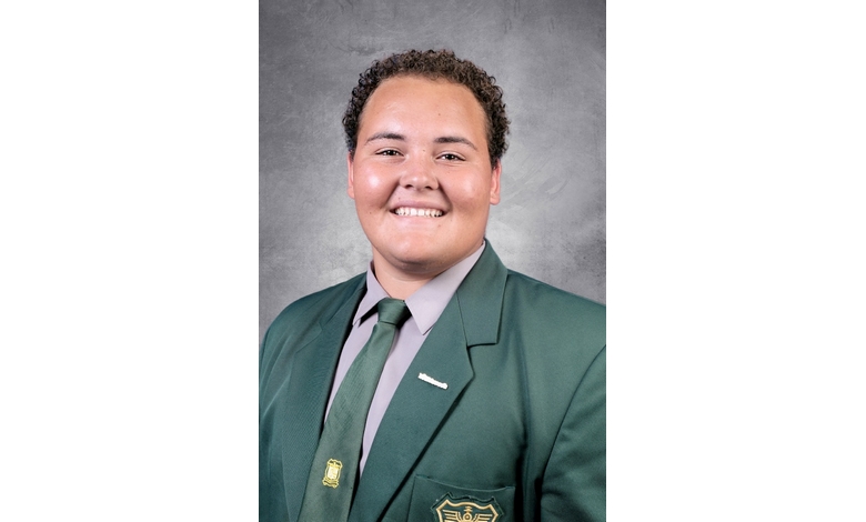 Smiling individual in a green blazer and tie against a gray background. The expression is friendly and confident. Emblem on blazer pocket.
