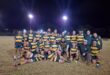A rugby team poses for a group photo on a field at night, under bright floodlights. Players wear matching green and yellow striped jerseys, smiling broadly.