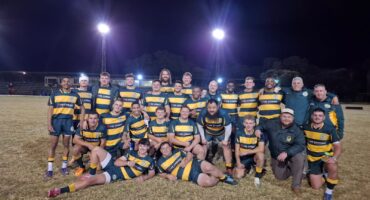 A rugby team poses for a group photo on a field at night, under bright floodlights. Players wear matching green and yellow striped jerseys, smiling broadly.
