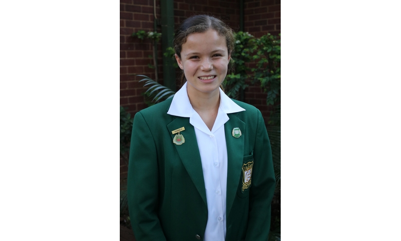 Smiling student wearing a green blazer with badges, white shirt, and standing outdoors against a brick wall and greenery, exuding confidence.