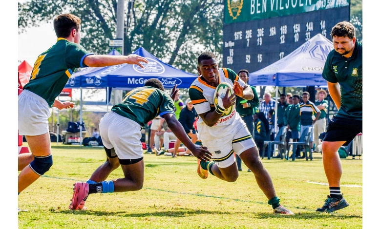 A rugby player in green and white runs with the ball, pursued by opposing players on a sunny field. Tents and a scoreboard are in the background.