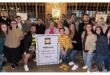 A group of 20 people gathered indoors for a photo, smiling warmly. They hold a large sign that reads "Welkom! Matriek 2016, 10 Jaar Reünie, 30 Mei 2026." The setting is casual and joyful, suggesting a reunion.