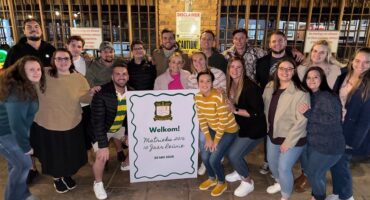 A group of 20 people gathered indoors for a photo, smiling warmly. They hold a large sign that reads "Welkom! Matriek 2016, 10 Jaar Reünie, 30 Mei 2026." The setting is casual and joyful, suggesting a reunion.