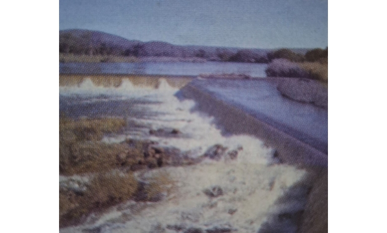 A river flows over a dam, creating a cascading waterfall. Surrounding hills and trees line the horizon under a clear blue sky, evoking tranquility.