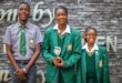 Three students stand smiling in front of a stone wall; two girls in green uniforms hold a trophy, and a boy in a gray shirt and green tie stands beside them.