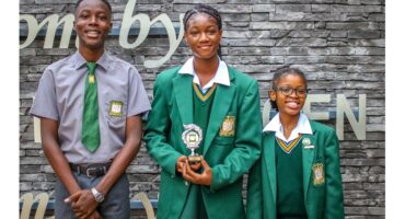 Three students stand smiling in front of a stone wall; two girls in green uniforms hold a trophy, and a boy in a gray shirt and green tie stands beside them.