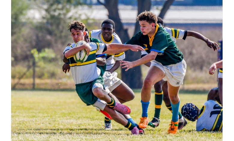 Boys in colorful uniforms engaged in a dynamic rugby tackle on a grassy field. Intense expressions show focus and determination under a clear sky.