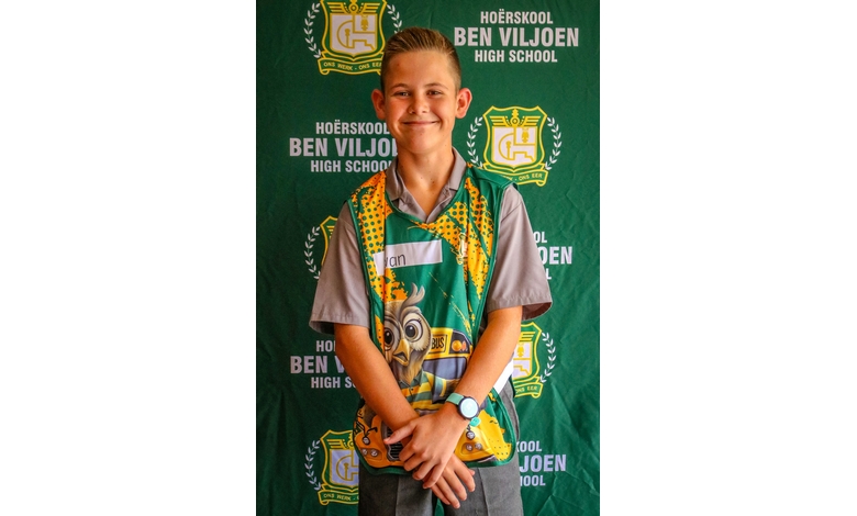 Smiling boy stands in front of a green "Hoërskool Ben Viljoen High School" backdrop. He wears a colorful vest and grey shirt, exuding a cheerful vibe.