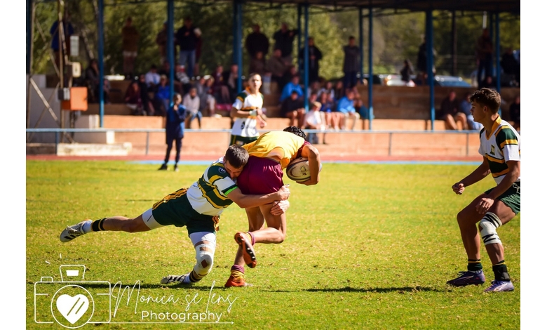 A rugby player in a yellow jersey is tackled by an opponent in green and white, on a sunny field. Spectators watch from the stands in the background.
