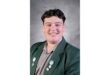 Smiling person in a green blazer with several pins on the lapel, set against a gray textured background. The image conveys a positive and friendly tone.