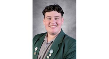 Smiling person in a green blazer with several pins on the lapel, set against a gray textured background. The image conveys a positive and friendly tone.