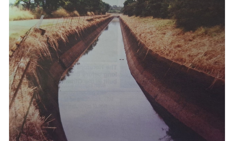 A narrow, concrete irrigation canal runs through a rural landscape. Dry grass lines the canal's banks under clear skies, suggesting a serene atmosphere.