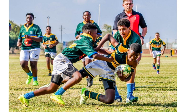 A group of young rugby players in green and yellow jerseys intensely tackle another player, who is holding a ball, on a sunny field. A coach observes closely.