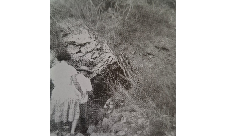 Black and white image of two people walking towards a rocky cave entrance surrounded by grass. The scene conveys curiosity and exploration.