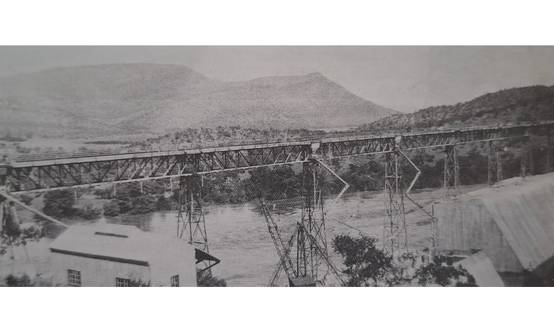 Black and white photo of a long, elevated bridge crossing a dam. Steel supports structure the bridge. Hills are visible in the background.