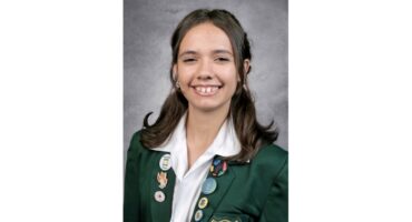 Smiling young person in a green blazer adorned with various colorful pins and badges. The background is a neutral gray, conveying a formal tone.