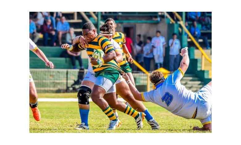 Rugby players in action; a player in green and yellow dodges a tackle from an opposing player in white and blue. Dynamic and intense scene.