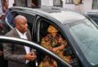 A woman wearing a colorful outfit and hat sits in a car, smiling and holding the steering wheel. A man in a suit stands beside the door, gesturing warmly.