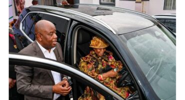 A woman wearing a colorful outfit and hat sits in a car, smiling and holding the steering wheel. A man in a suit stands beside the door, gesturing warmly.