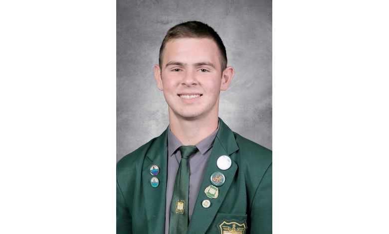 Young person smiling in a green blazer adorned with multiple pins and a badge, against a gray textured backdrop. The mood is confident and professional.