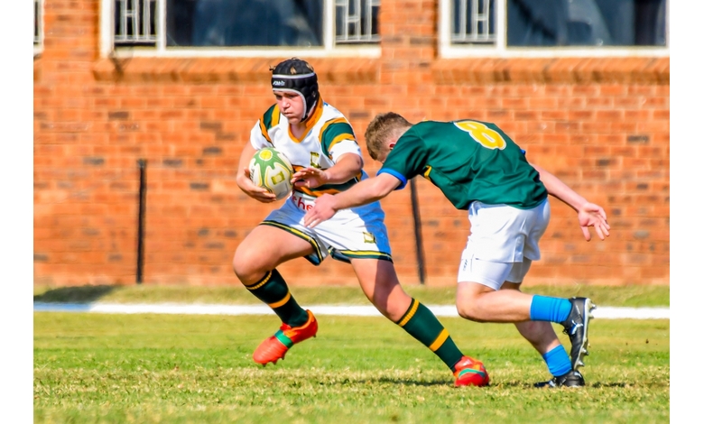 Two rugby players in action on a grassy field. One, wearing a helmet and holding the ball, evades a tackle. Vibrant jerseys, brick building backdrop.