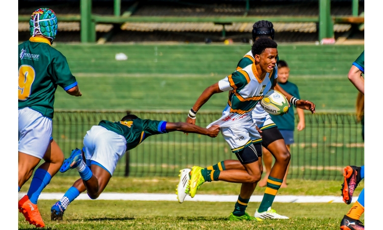 A rugby player in a green and white uniform sprints forward, holding the ball, as an opponent in green dives to tackle him on a sunny field.