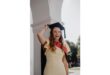 A joyful woman in a sleeveless yellow dress and graduation cap stands under an arch, smiling broadly. Sunlight filters through nearby trees, creating a celebratory mood.