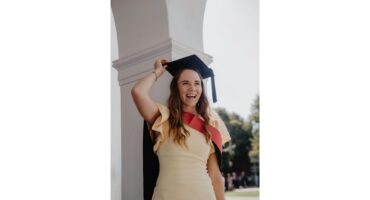 A joyful woman in a sleeveless yellow dress and graduation cap stands under an arch, smiling broadly. Sunlight filters through nearby trees, creating a celebratory mood.