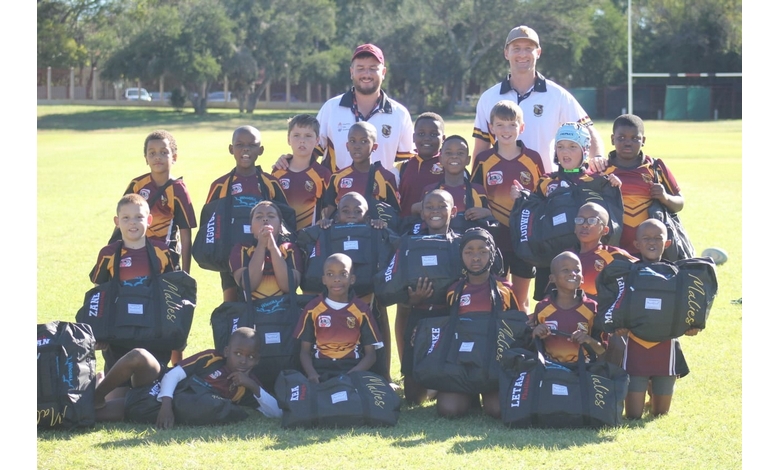 A youth rugby team poses on a sunny field, wearing maroon and yellow jerseys. Two adult coaches stand behind the smiling kids holding gear bags.