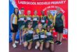 A girls' sports team poses in green uniforms holding certificates, smiling with two coaches on either side. Background features a red school banner.