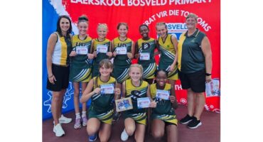 A girls' sports team poses in green uniforms holding certificates, smiling with two coaches on either side. Background features a red school banner.