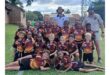 A junior rugby team poses on a grassy field with two adults. The boys wear matching maroon and yellow jerseys, smiling happily in a group.