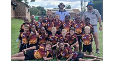 A junior rugby team poses on a grassy field with two adults. The boys wear matching maroon and yellow jerseys, smiling happily in a group.