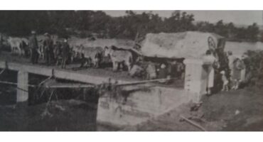 Black and white photo of a large mule-drawn cart crossing a narrow bridge, with people and animals nearby. The setting appears rural and historical.