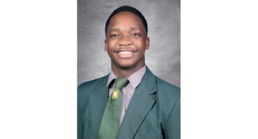 Smiling young man in a green school blazer and tie against a gray background, projecting confidence and positivity.