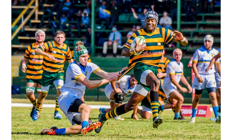 A rugby player in a green and yellow striped jersey runs with the ball, evading a tackle from an opposing player in a white and blue uniform. The scene is action-packed and intense, conveying a sense of urgency and competition on a grassy field with spectators in the background.
