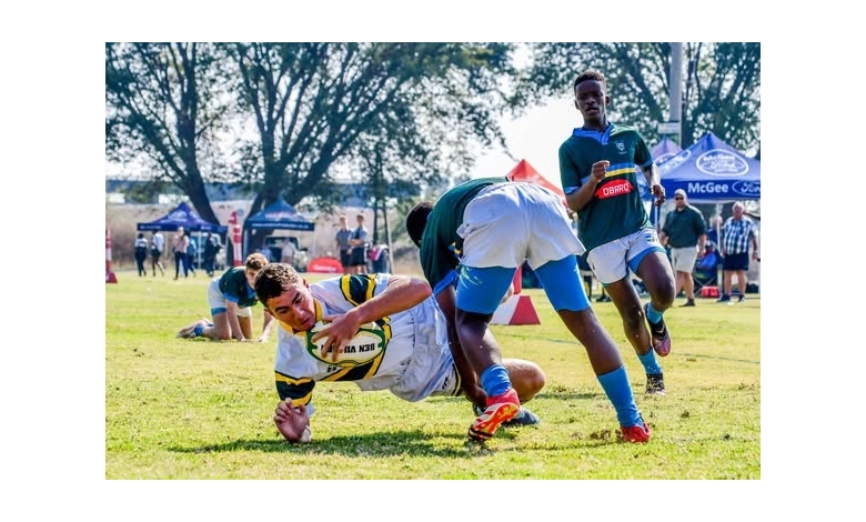 A rugby player in a white and yellow jersey dives to the ground clutching the ball, as two opponents in green tackle him. The scene is dynamic, on a sunlit field with spectators and trees in the background, conveying action and intensity.